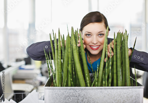 Photo : Smiling businesswoman peering from behind plant in office Smiling businesswoman peering from behind plant in office