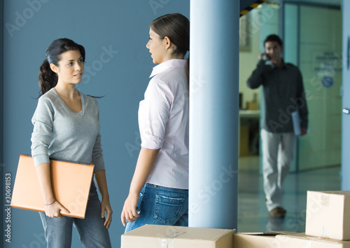 Foto: Two young women standing face to face talking, man in background using cell phone Two young women standing face to face talking, man in background using cell phone