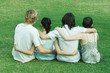 Four young friends sitting on grass, side by side, with arms around each other, rear view