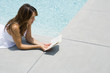 Woman lying on stomach next to pool, reading book