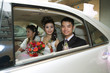 Bride and groom in car with bridesmaid, smiling at camera