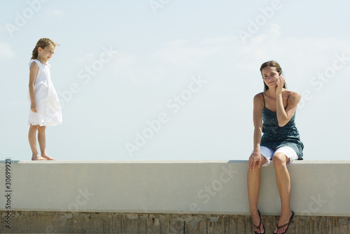 Photo: Young woman sitting on low wall, using cell phone, little girl standing nearby, both smiling Young woman sitting on low wall, using cell phone, little girl standing nearby, both smiling