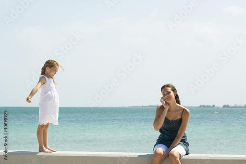 Photo: Little girl standing on low wall, watching older sister using cell phone Little girl standing on low wall, watching older sister using cell phone