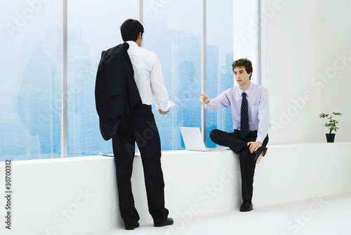 Photo: Two businessmen facing each other, one sitting on ledge, one standing Two businessmen facing each other, one sitting on ledge, one standing