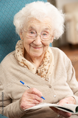 Foto: Senior Woman Relaxing In Chair At Home Completing Crossword Senior Woman Relaxing In Chair At Home Completing Crossword
