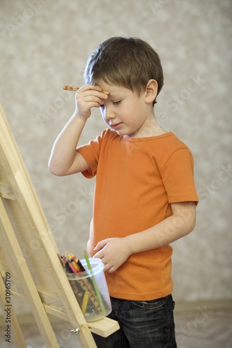 Photo: A young boy holding his hand with a pencil in up to his face, standing infront of a canvas with a pot of coloured pencils A young boy holding his hand with a pencil in up to his face, standing infront of a canvas with a pot of coloured pencils
