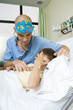 Boy lying in hospital bed holding head, doctor listening to boy with stethoscope