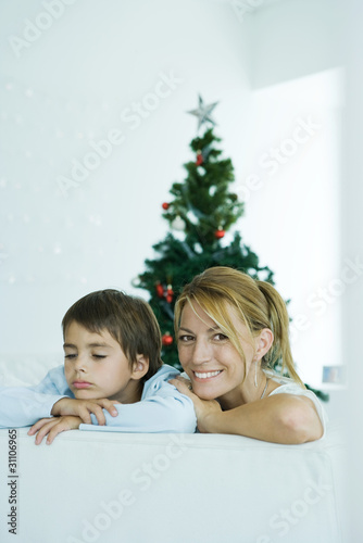 Photo: Boy and mother on sofa, Christmas tree in background, woman smiling at camera, boy frowning Boy and mother on sofa, Christmas tree in background, woman smiling at camera, boy frowning