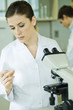 Female lab worker standing next to microscope, holding up and inspecting slide