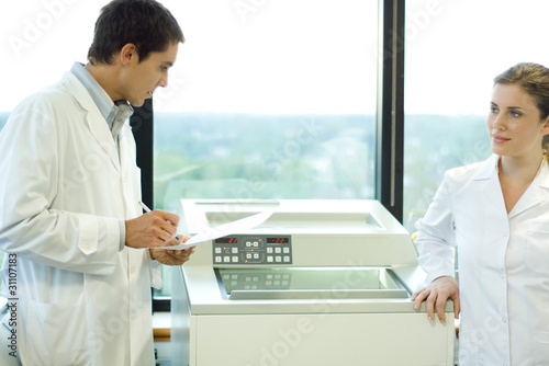 Photo: Male and female lab workers standing next to lab machine Male and female lab workers standing next to lab machine