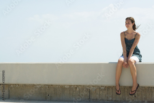 Photo: Young woman sitting on low wall outdoors, smiling, looking away Young woman sitting on low wall outdoors, smiling, looking away