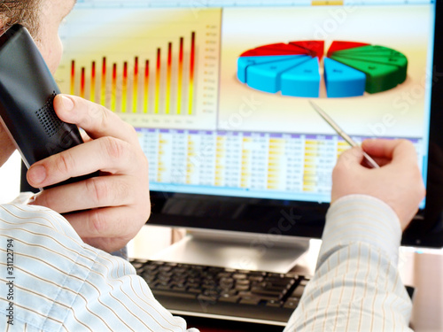 Photo: Man on a phone analizing financial data and charts on computer Man on a phone analizing financial data and charts on computer