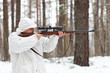 Sniper in white camouflage aiming with rifle at winter forest.