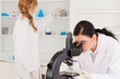 Dark-haired scientist looking through a microscope with her assi