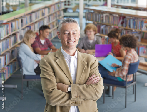 Photo: Smiling teacher standing in school library with study group on background Smiling teacher standing in school library with study group on background