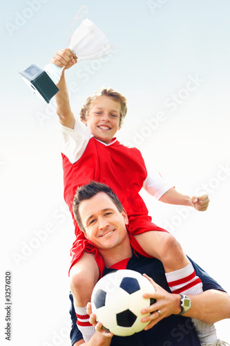 Foto: Soccer kid holds trophy while sitting over coach's shoulders Soccer kid holds trophy while sitting over coach's shoulders
