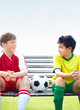 Two rival soccer players with a ball sitting together on bench