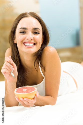 Photo: Smiling beautiful woman eating grapefruit on bed, at home Smiling beautiful woman eating grapefruit on bed, at home