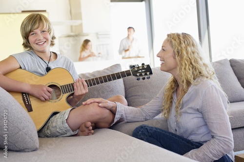 Photo: Teenage boy playing a guitar with his mother sitting near him and smiling Teenage boy playing a guitar with his mother sitting near him and smiling