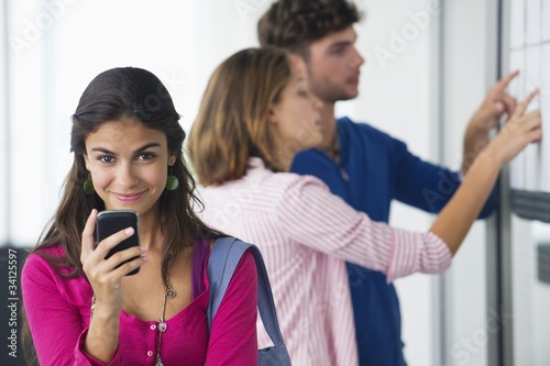 Foto: Portrait of university students using a mobile phone while friends checking for test results in bulletin board Portrait of university students using a mobile phone while friends checking for test results in bulletin board