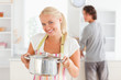 Woman posing with a boiler while her fiance is washing the dishe