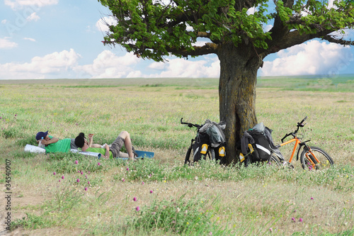 Photo: bicycle tourists resting near tree, field and blue sky bicycle tourists resting near tree, field and blue sky