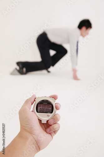 Photo: Human hand holding stopwatch with businessman crouching on starting block in the background Human hand holding stopwatch with businessman crouching on starting block in the background