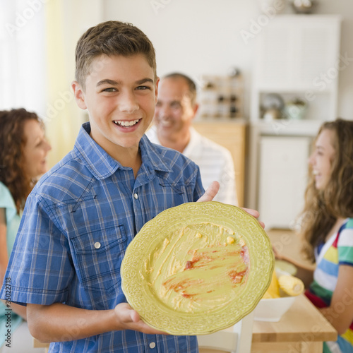 Photo: Smiling boy displaying empty plate with family at table in background Smiling boy displaying empty plate with family at table in background