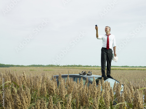 Photo: Caucasian businessman on top of car in field looking at gps device Caucasian businessman on top of car in field looking at gps device