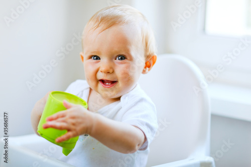Photo: Cheerful baby girl sitting in chair and holding baby cup Cheerful baby girl sitting in chair and holding baby cup