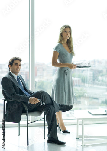 Photo: Office lobby, man sitting while woman stands in background holding magazine Office lobby, man sitting while woman stands in background holding magazine