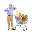 Shocked senior looking at store receipt next to a shopping cart