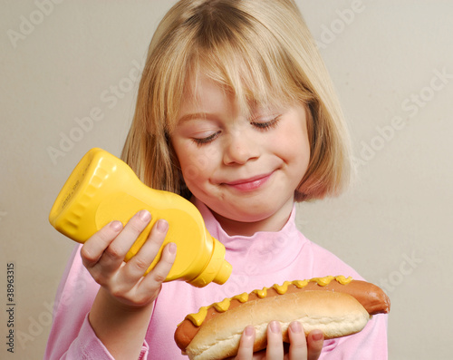 Photo: Niña feliz comiendo perro caliente,Hot Dog.Colocando mostaza. Niña feliz comiendo perro caliente,Hot Dog.Colocando mostaza.