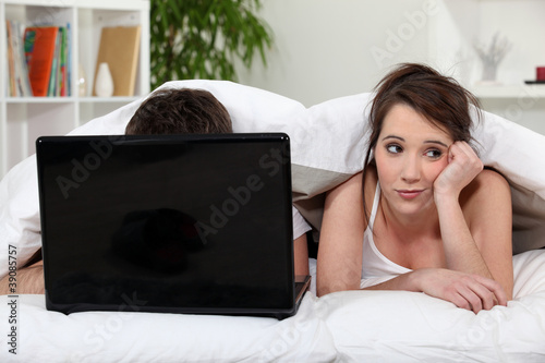 Photo: Unimpressed woman in bed with her partner and his laptop Unimpressed woman in bed with her partner and his laptop