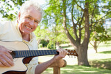 Photo: Man smiling while playing a guitar while sitting on a bench with a tree in the back Man smiling while playing a guitar while sitting on a bench with a tree in the back