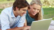 A smiling couple lie in the park with a laptop as they look up into the camera