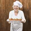 Foto: senior woman cook holding a tray with muffins against a wooden b senior woman cook holding a tray with muffins against a wooden b