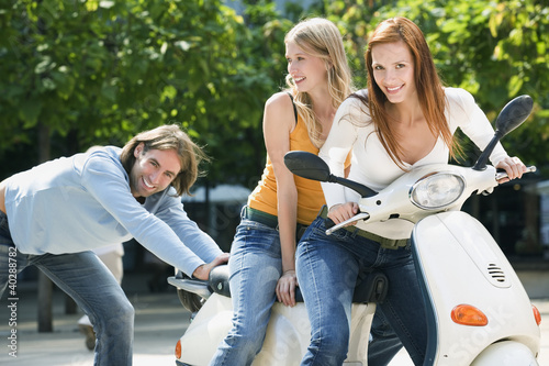 Photo: Young man pushing young women on motor scooter, smiling Young man pushing young women on motor scooter, smiling