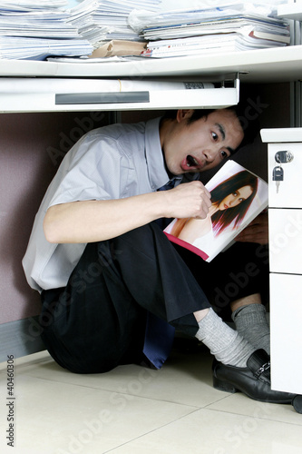 Foto: Man hiding under his desk reading pornographic magazine. Man hiding under his desk reading pornographic magazine.