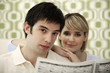 Man and woman smiling at camera while reading newspaper