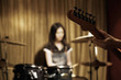 Focus on hand playing guitar, young woman playing drums in the background