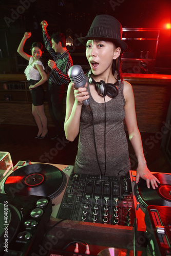 Photo: Woman spinning turntable while singing with people dancing in the background Woman spinning turntable while singing with people dancing in the background
