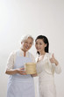 Photo : Senior woman with apron holding bamboo steamers, woman posing beside her showing thumbs up Senior woman with apron holding bamboo steamers, woman posing beside her showing thumbs up