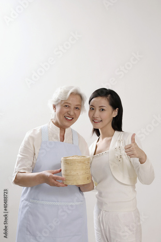Photo: Senior woman with apron holding bamboo steamers, woman posing beside her showing thumbs up Senior woman with apron holding bamboo steamers, woman posing beside her showing thumbs up