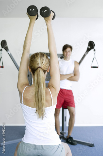 Foto: Woman lifting dumbells with a man watching her in the background Woman lifting dumbells with a man watching her in the background