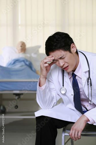 Photo: Doctor having headache, dummy patient on the bed in the background Doctor having headache, dummy patient on the bed in the background