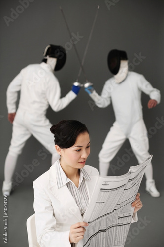 Photo: Businesswoman reading newspaper with two men fencing in the background Businesswoman reading newspaper with two men fencing in the background