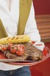 Foto: Woman holding T-bone steak, corn on the cob & cherry tomatoes Woman holding T-bone steak, corn on the cob & cherry tomatoes
