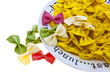 Uncooked rainbow farfalle pasta spilling from plate on white bac Uncooked rainbow farfalle pasta spilling from plate on white bac