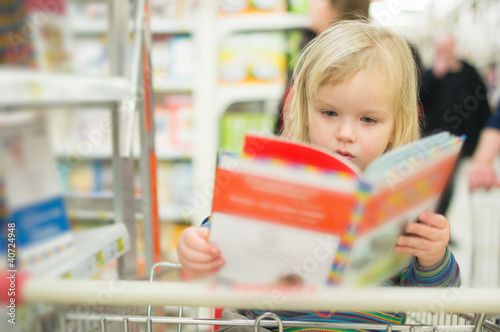 Photo: Adorable baby with book near book stand in supermarket Adorable baby with book near book stand in supermarket